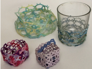 Four pieces of 3D tatting, one is placed around a glass, one is coloured green and yellow, another is in shades of pink