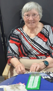Woman with grey hair, glassess, wearing red, black and white shirt, is smiling and making bobbin lace