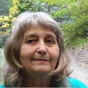 Woman with shoulder length grey hair, standing in front of several trees and a brick wall, wearing green top