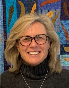 Smiling woman is standing in front of colourful quilt, shoulder length blonde hair, wearing glasses, grey turtle neck