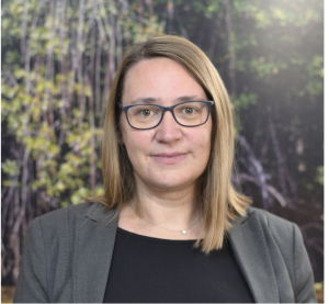Photo of woman with shoulder length light brown hair, glasses, smiling, wearing black top and grey jacket with vines in the background