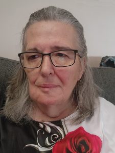 Woman with grey shoulder length hair, half pulled back, wearing glasses and white and black top with a red rose