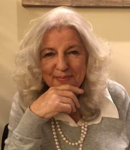 Woman with shoulder length grey hair, wearing grey top, white blouse and pearls, smiling, looking into camera while leaning on hand