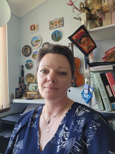 Woman with short brown hair, wearing a black and white patterned top, standing in front of decorative wall plates and book shelf
