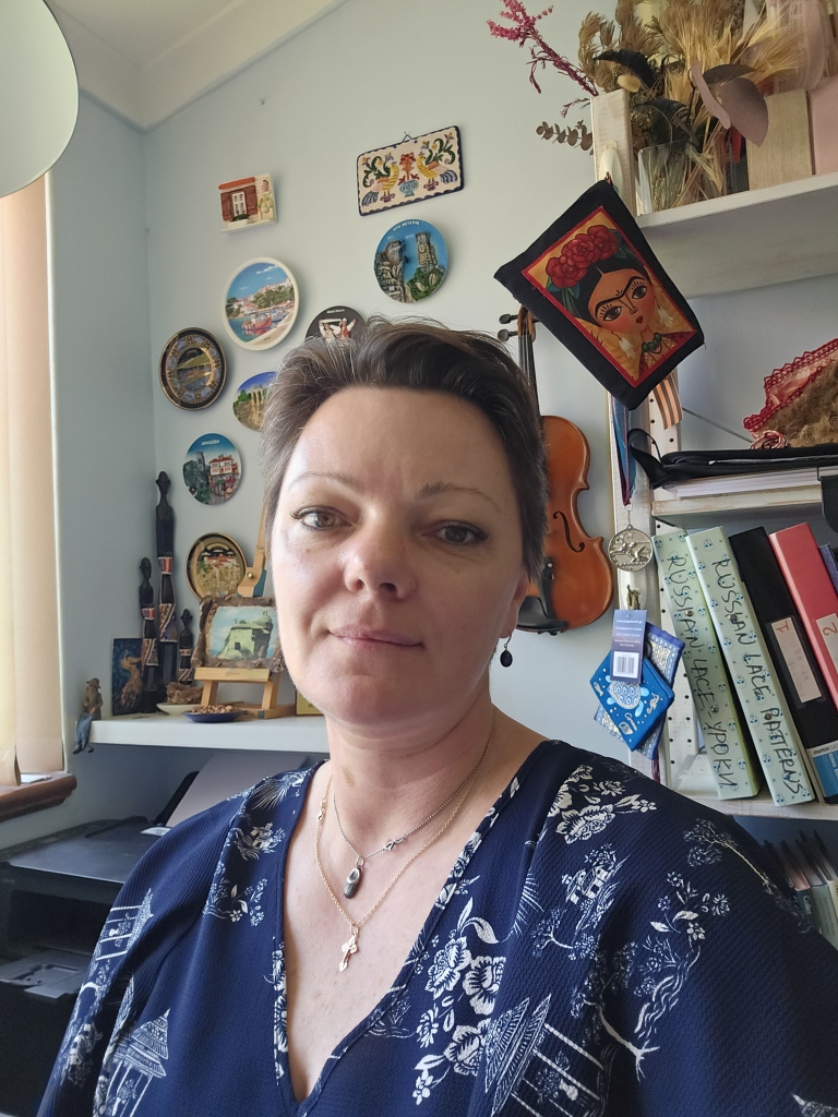 Woman with short brown hair, wearing a navy blouse with white patterns, standing in front of a bookshelf